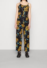 Černý overálek s žlutým květinovým vzorem a zelenými akcenty, s nastavitelnými špagetovými ramínky a knoflíkovými detaily vpředu.