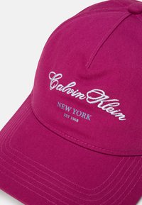 Fuksiaroosa pesapallikeeper, mis on valmistatud tekstuuritud kangast, millel on tikitud "Calvin Klein" logo ja "NEW YORK EST 1968" valges ja helesinises värvitoonis.