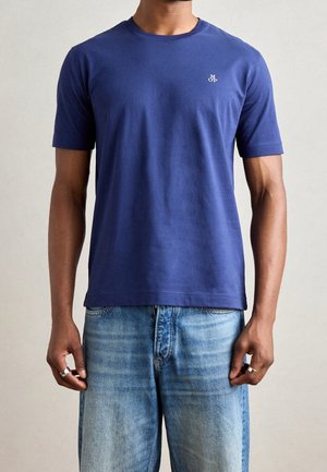 Hombre con camiseta azul marino lisa de manga corta con pequeño logo blanco en el pecho, combinada con jeans de mezclilla azul claro y anillos plateados.