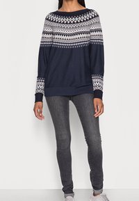 Maglione blu marina con motivi geometrici bianchi, scollatura rotonda e maniche lunghe; abbinato a jeans scuri attillati e scarpe chiare.