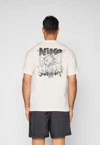 Cremefarvet bomulds t-shirt med et stort sort grafisk tryk af en basketball og "Nike Hoops" tekst på ryggen. Korte ærmer, afslappet pasform.