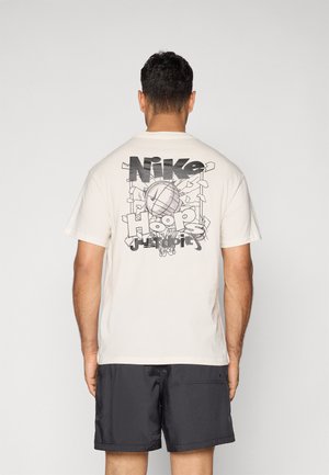 Cremefarvet bomulds t-shirt med et stort sort grafisk tryk af en basketball og "Nike Hoops" tekst på ryggen. Korte ærmer, afslappet pasform.