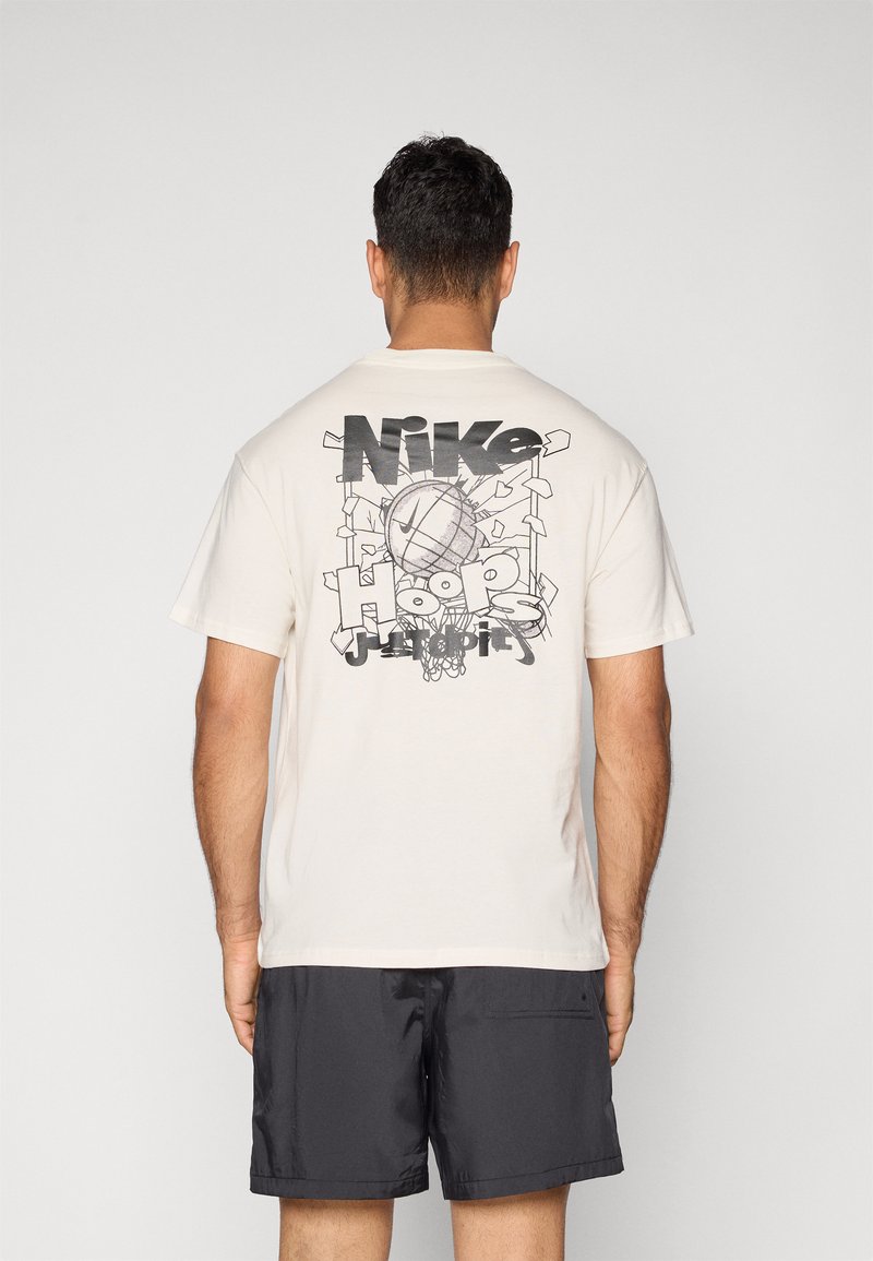 Cremefarvet bomulds t-shirt med et stort sort grafisk tryk af en basketball og "Nike Hoops" tekst på ryggen. Korte ærmer, afslappet pasform.