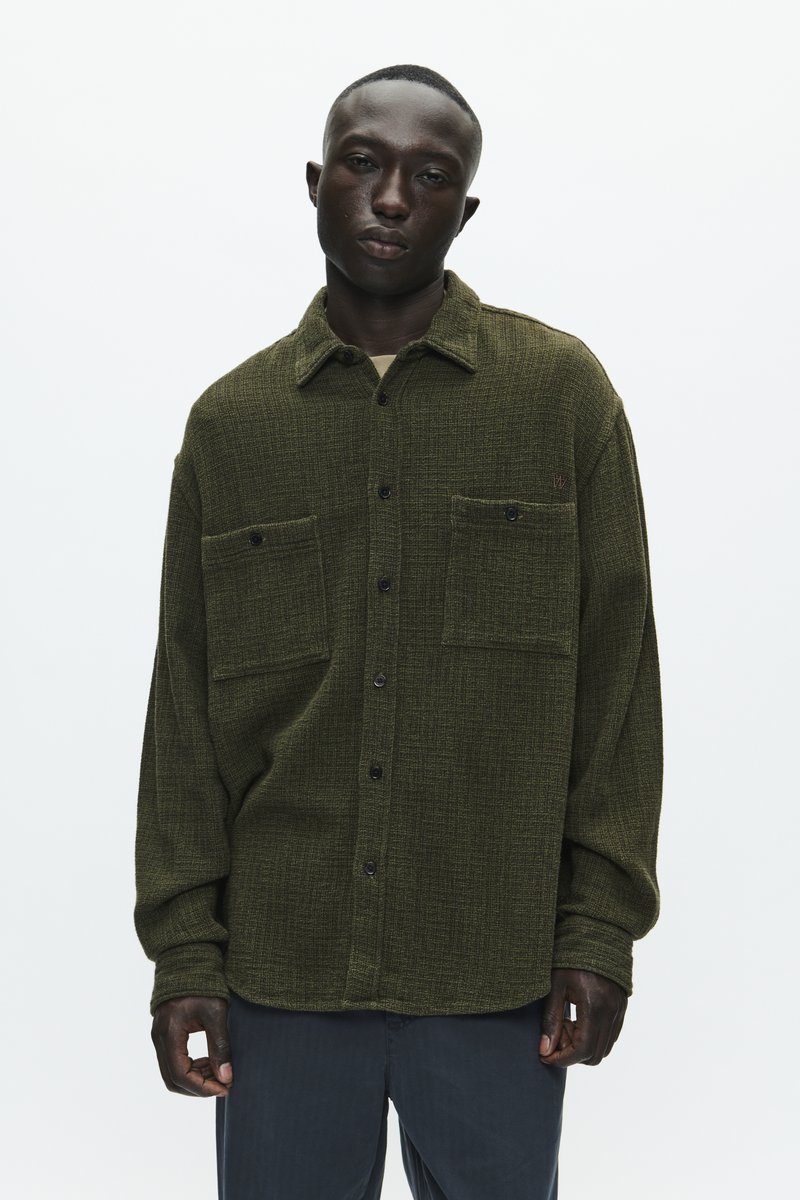 Olijfgroene, gestructureerde shirt met een kraaghalslijn, twee borstzakken en een knoopsluiting aan de voorkant, gecombineerd met donkere broeken.