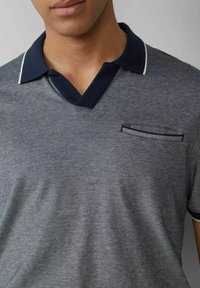 Graues Poloshirt mit navy-blauem Kragen und weißen Akzenten, ausgestattet mit einer Brusttasche, die einen gestreiften Akzent und strukturierten Stoff aufweist.