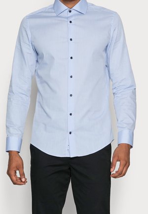 Mężczyzna ubrany w dopasowaną, jasnoniebieską koszulę z długim rękawem, ciemnymi guzikami i czarne spodnie, stojący na jednolitym tle.