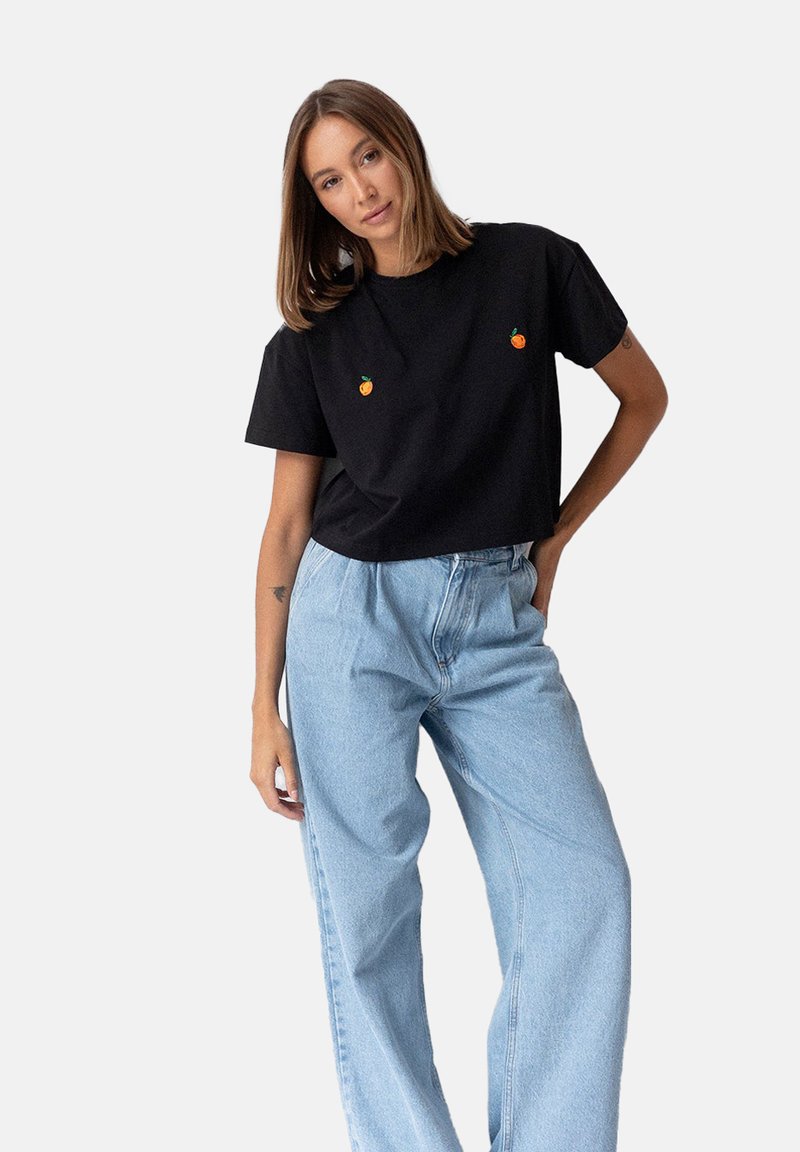 Czarna bawełniana koszulka z krótkimi rękawami, ozdobiona dwoma małymi haftami w kształcie pomarańczowych owoców. Noszona z jasnoniebieskimi szerokimi spodniami.