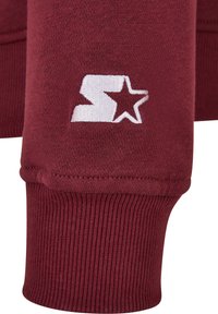 Burgundi színű pulóver bordás mandzsettával, fehér hímzett logóval és csillag részlettel. Puha szövet textúrával, sportos dizájnnal.