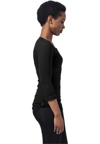Haut noir à manches longues avec texture côtelée, coupe slim et manches roulées. Porté sur des leggings noirs, avec un ourlet asymétrique.