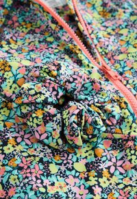 Blumenstoff mit pinken, gelben, grünen und schwarzen Blumen, ausgestattet mit einem passenden Scrunchie und einem korallenfarbigen Reißverschlussdetail.