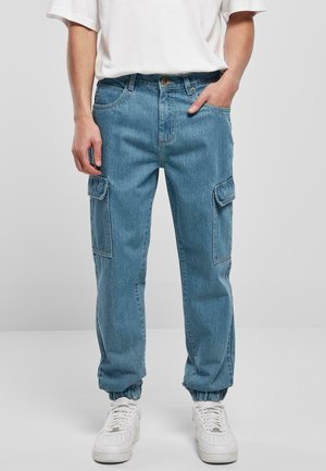 Hombre con pantalones cargo de denim azul claro con puños elásticos en los tobillos, zapatillas blancas y una camiseta blanca de manga corta, mano en el bolsillo.