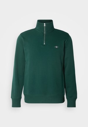 Grønn glidelåsgenser. Har høy krage, lange ermer, og en liten logo på brystet. Mykt, glatt stoff.