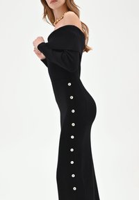 Schwarzes, geripptes, schulterfreies Kleid mit figurbetonter Silhouette und seitlichen Knopfverzierungen entlang der linken Naht. Glatte Textur.