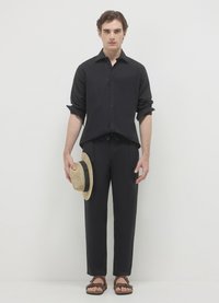 Schwarzes Hemd mit Knöpfen und passende Hose, beide aus leichtem Stoff. Das Modell hält einen Strohhut und trägt braune Sandalen.