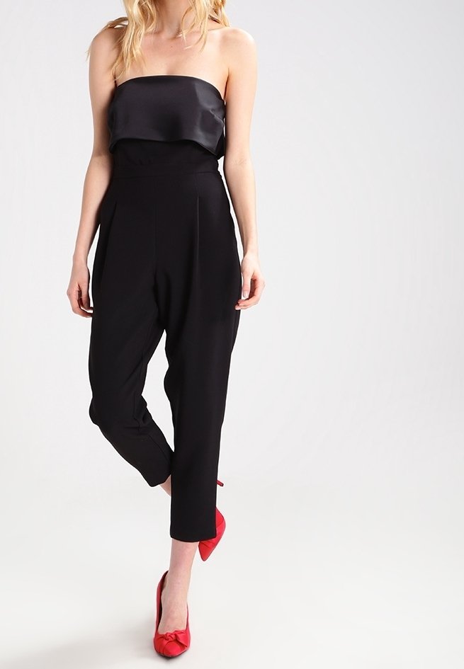 Vrouw in een strapless zwarte jumpsuit en rode hakken, staande met één been gekruist voor het andere tegen een effen achtergrond.