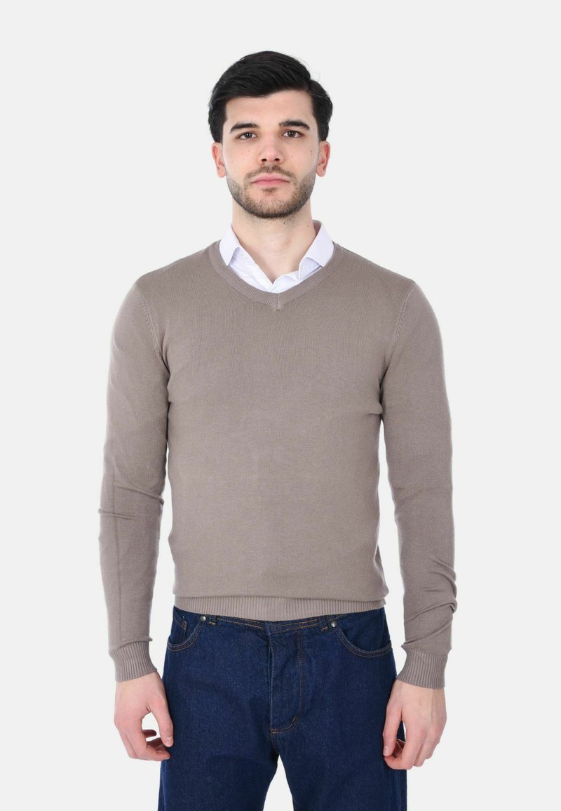 Uomo con capelli corti e scuri che indossa un maglione beige a scollo a V sopra una camicia bianca e jeans blu, in piedi contro uno sfondo chiaro e semplice.