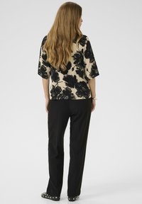 Moters su ilgais rudais plaukais, vilkinčios kreminę bluzą su dideliu juodu gėlių raštu ir juodomis kelnėmis, stovinčios atsisukusios atgal ant vientiso fono.