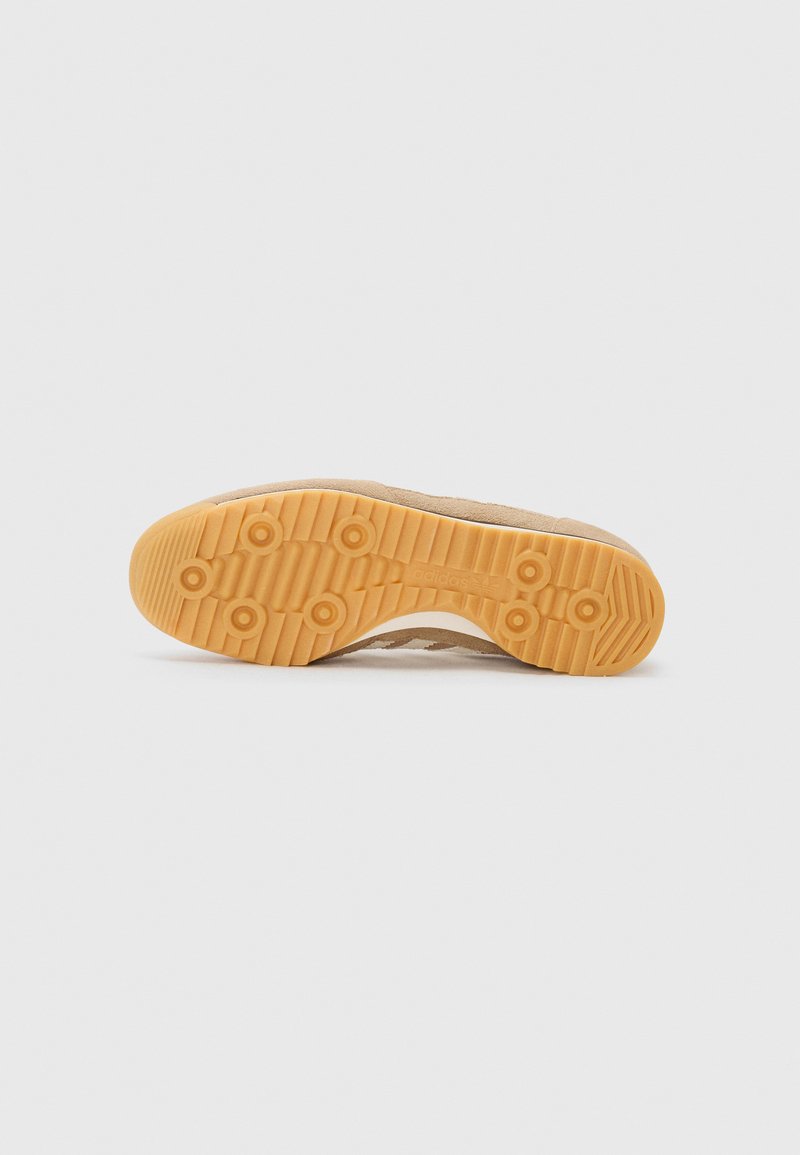 Zapatilla con parte superior de ante en tono beige y suela de goma con agarres circulares. El diseño incluye un acabado texturizado y un branding sutil.