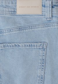 Poche arrière de jean en denim bleu clair avec passant de ceinture et étiquette en cuir blanc cousue, embossée du texte « Street One Studio ».