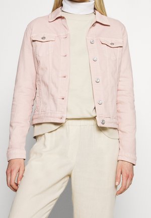 Mujer con chaqueta vaquera de botones rosa claro sobre jersey de cuello alto crema y pantalones crema a juego, posando frente a un fondo blanco.