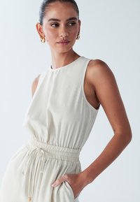 Bež brezrokavnik z nabranim pasom, bočnima žepoma in gladko teksturo na svetli podlagi. Viden je zlat obročni uhan.