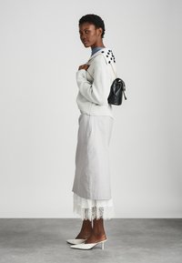 Ljusgrå tröja med strukturerad stickning, lagd över en blå topp. Lång grå kjol med spetsdetalj, matchad med vita klackskor och en svart väska.