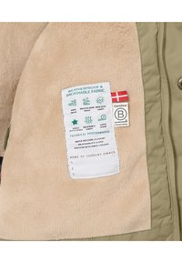 Doublure en fausse fourrure beige avec un extérieur en tissu vert. Dispose d'étiquettes d'entretien et de performance, y compris des détails sur le tissu imperméable et respirant.