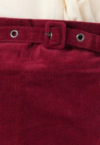 Zbliżenie bordowej plisowanej spódnicy sztruksowej z pasującym paskiem z tkaniny i srebrnymi oczkami na talii.