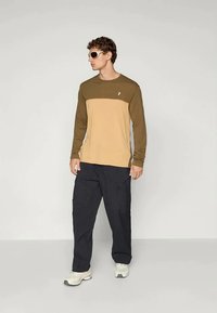 Long-sleeve top featuring brown and tan color-block design, paired with dark cargo pants; model wears sunglasses and sneakers.
