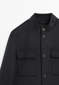 Giacca nera in misto lana con colletto alto, due tasche sul petto e cinque bottoni. Presenta una texture liscia e una vestibilità sartoriale.