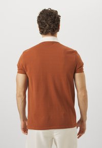 Trumpas rankovėmis polo marškinėliai rusvai oranžinės spalvos, su kontrastinga balta apykakle, pagaminti iš lengvos medžiagos su lygiu paviršiumi.