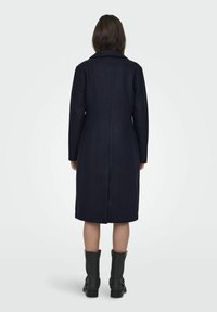 Lungo cappotto blu navy realizzato in un misto lana, con colletto a rever, silhouette dritta e lunghezza fino al polpaccio, abbinato a stivali neri alla caviglia.