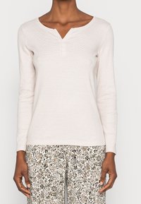 Helles rosa langärmliges Henley-Oberteil mit V-Ausschnitt und drei Knöpfen, kombiniert mit einer floral gemusterten Hose in neutralen Tönen. Weicher Stoff, figurbetontes Design.