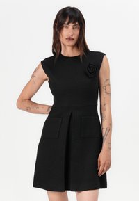 Black sleeveless dress with textured fabric, featuring a floral accent and two front pockets, flared skirt, and slight pleat detail.