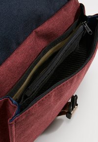 Vinröd tygpåse med marinblå detaljer, med ett dragkedjefack och en svart nätficka inuti. Inkluderar ett spänne och robust sömnad.