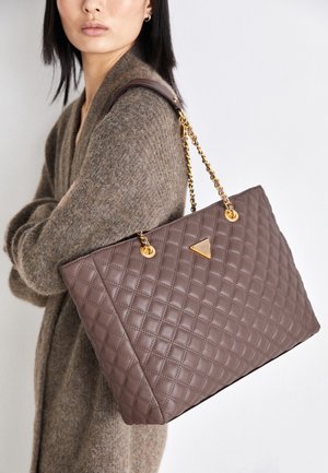 Femme portant un cardigan en maille marron, portant un grand sac à main matelassé marron avec des bandoulières en chaîne dorée sur l’épaule.