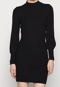 Robe mini en maille côtelée noire avec manches longues bouffantes et col rond montant, silhouette ajustée sur une personne debout.