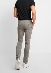 Man som bär grå slim-fit byxor, svart t-shirt och vita sneakers, stående med en hand i fickan mot en enfärgad bakgrund.