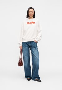 Bela oversized majica z rdečim srčnim vzorcem z napisom "HUGO", kombinirana z modrimi širokimi kavbojkami in majhno rjavo torbico. Črni balerinki.