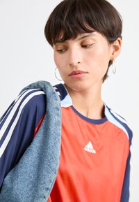 Raudona Adidas sportinė marškinė su tamsiai mėlynomis rankovėmis, puikuojasi baltais trimis dryžiais ir apvaliu kaklu. Šviesiai mėlynos akcentai pečių srityje.