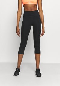 Kvinde iført sorte høj taljede cropped leggings, sorte løbesko og en neongul sports-bh, der står foran en ensfarvet hvid baggrund.