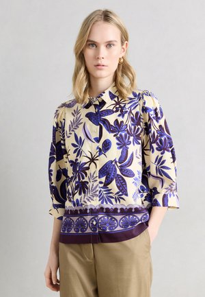 Jeune femme aux cheveux blonds portant une chemise beige à boutons avec des motifs floraux bleus et violets et un pantalon beige, debout devant un fond clair.