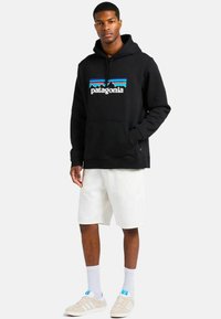 Schwarzer Baumwoll-Hoodie mit Fronttasche, ausgestattet mit einem blauen und weißen "Patagonia"-Logo und einem Berggrafik. Getragen mit weißen Shorts und beigen Turnschuhen.