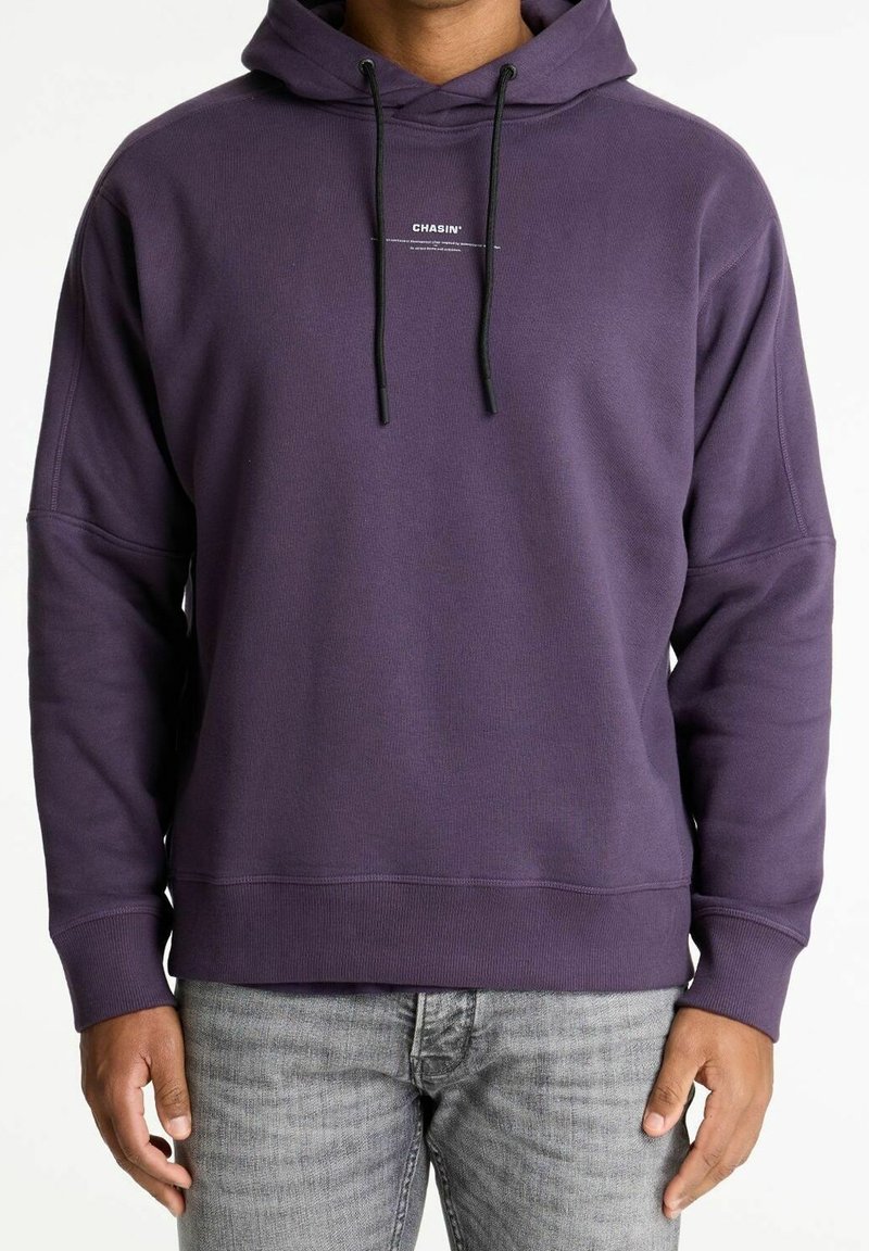 Homme portant un sweat à capuche violet foncé avec des cordons noirs et un petit texte blanc « CHASIN' » au centre de la poitrine, associé à un jean gris clair.