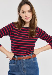 Langsleeve top in marineblauw met rode horizontale strepen, met een ronde halslijn. Gecombineerd met high-waisted blauwe spijkerbroeken en een leren bruine riem.