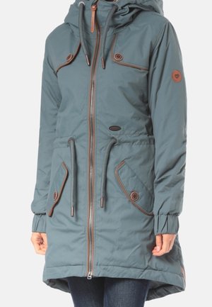 Long manteau d'hiver bleu sarcelle avec capuche, fermeture éclair à l'avant, cordons à la taille et à la capuche, poches à rabat boutonnées, porté sur un pantalon foncé.