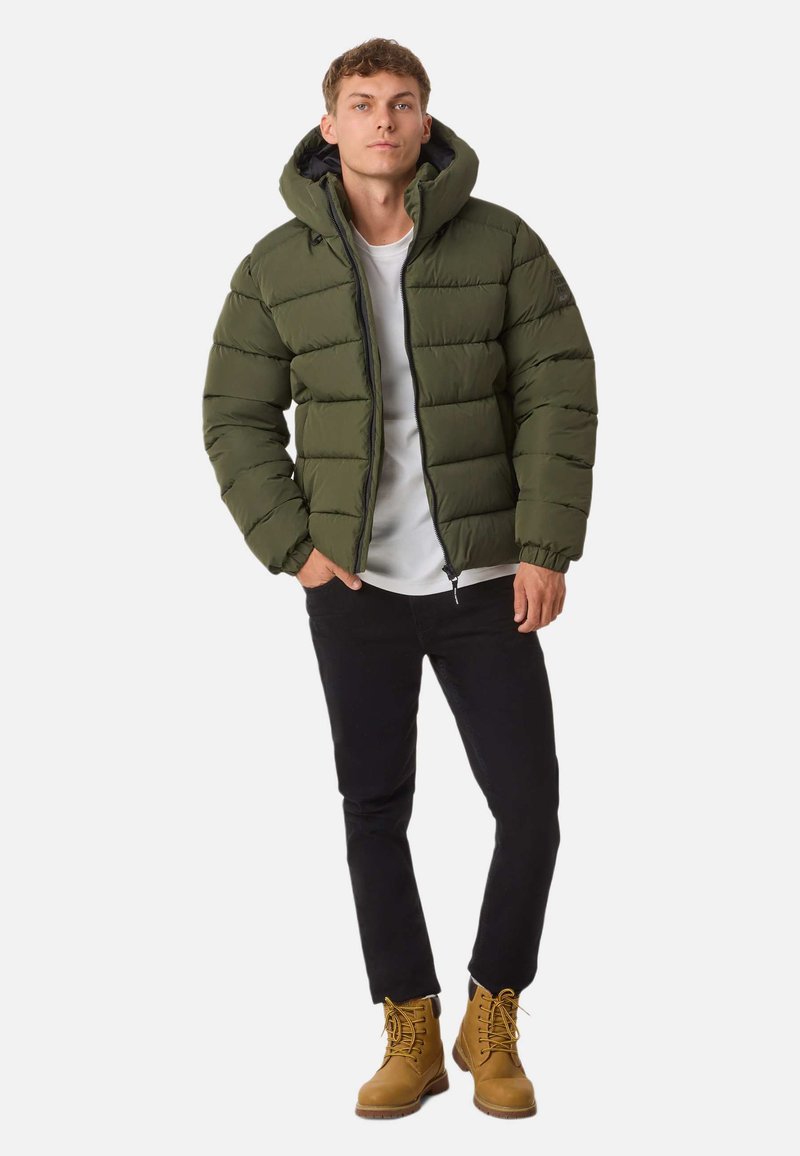 Zielona kurtka puchowa z kapturem, z pikowanymi sekcjami; noszona na białej koszuli i czarnych dżinsach. Beżowe buty z sznurówkami i gumowymi podeszwami.