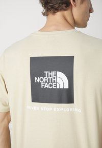 Homme portant un t-shirt beige avec un logo carré noir et le texte blanc "THE NORTH FACE" ainsi que le slogan "NEVER STOP EXPLORING" au dos.