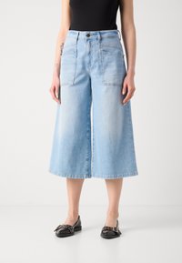 Žena v světlých modrých širokých zkrácených džínách s velkými předními kapsami a černými mokasínami se sponou, stojící na bílé podlaze.