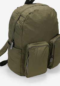 Mochila de nylon verde con forma redondeada, que cuenta con dos bolsillos frontales con cremallera y correas ajustables. El material tiene un acabado texturizado.